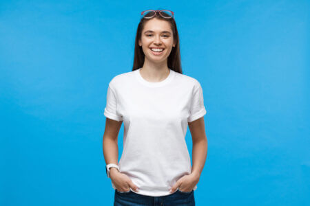 Young Female Standing With Hands In Pockets, Wearing Blank White T Shirt With Copy Space, Isolated On Blue Background